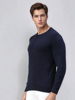 A man wears the 98degreenorth Men’s Dark Denim Merino Wool Sweater with white pants against a light grey background.