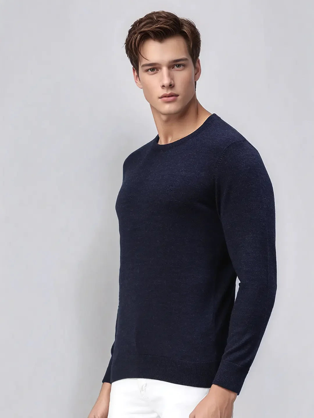 A man wears the 98degreenorth Men’s Dark Denim Merino Wool Sweater with white pants against a light grey background.