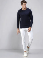 A man wears the 98degreenorth Men’s Dark Denim Merino Wool Sweater with white pants, standing by a light gray backdrop.