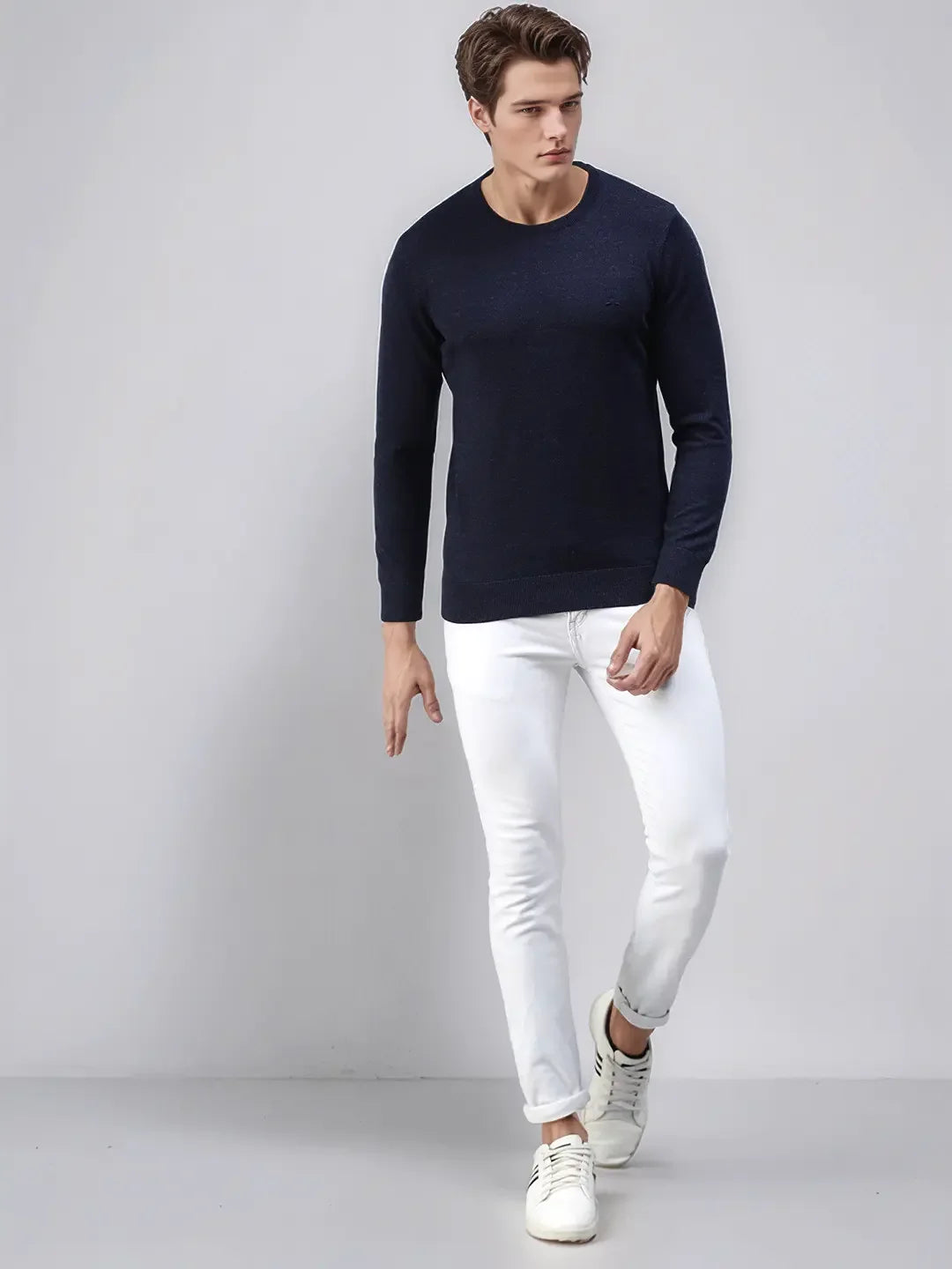 A man wears the 98degreenorth Men’s Dark Denim Merino Wool Sweater with white pants, standing by a light gray backdrop.