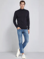 A man wears a 98degreenorth dark navy merino wool turtle neck sweater, blue jeans, and white sneakers.