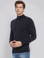 A man wears a 98degreenorth Men’s Dark Navy Merino Wool Turtle Neck Sweater and blue jeans against a light grey background.