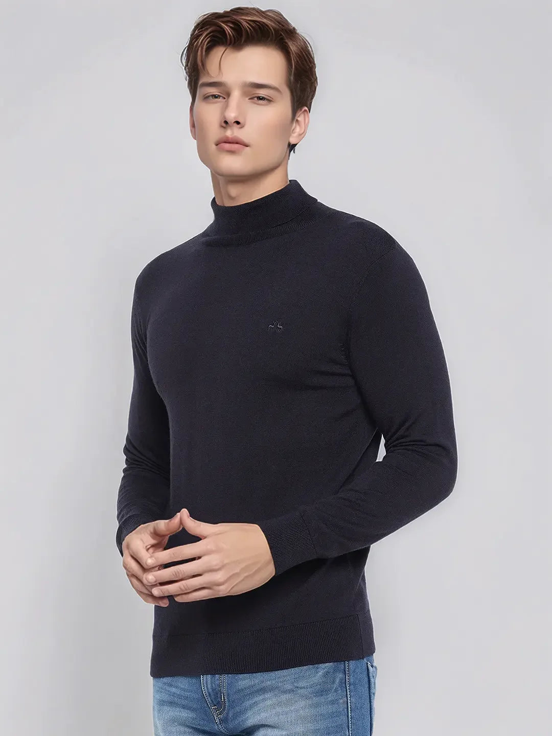 A man wears a 98degreenorth Men’s Dark Navy Merino Wool Turtle Neck Sweater and blue jeans against a light grey background.