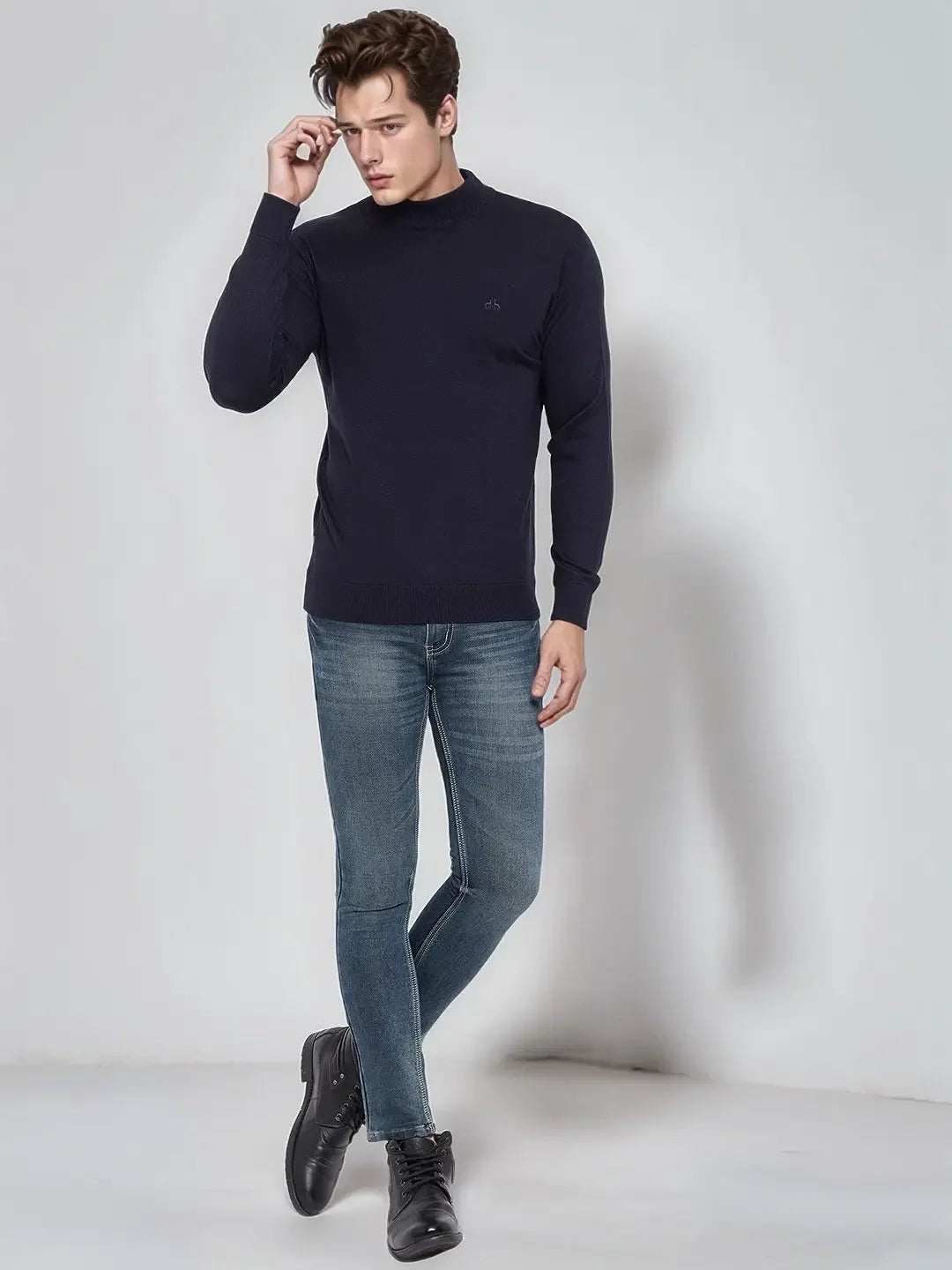 Man wearing 98degreenorth Dark Navy Merino Wool Turtle Neck Sweater, blue jeans, and black boots against a light wall.