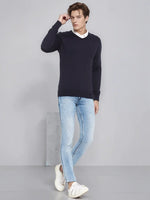 Man wears 98degreenorth Men’s Dark Navy Merino Wool V-Neck Sweater over a white shirt and jeans, standing by gray wall.