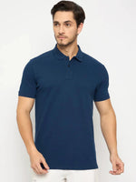 Man in 98degreenorth Men's Denim Blue Solid Cotton Pique Polo T-Shirt, paired with white pants against a plain background.