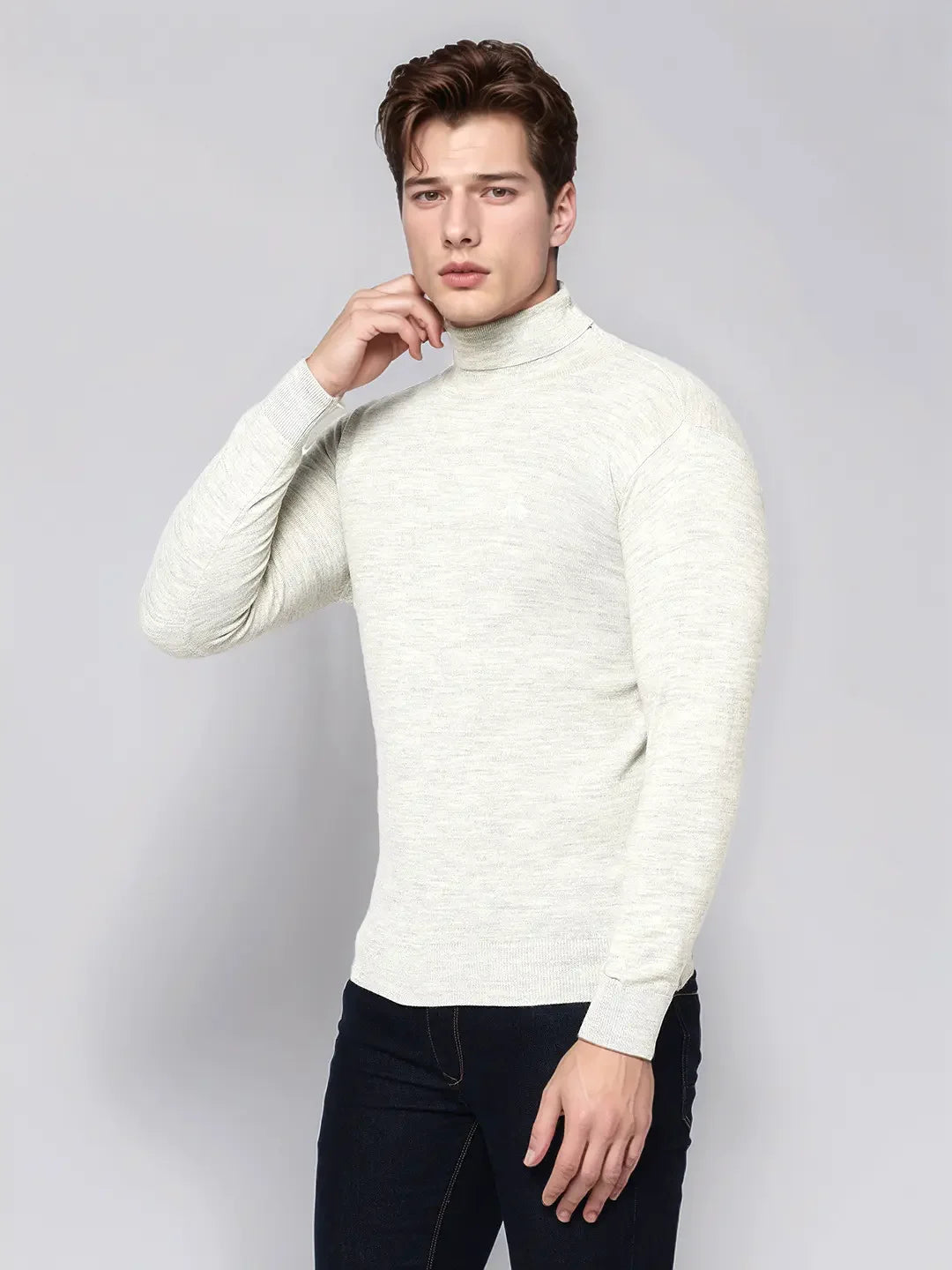 Man wears 98degreenorth Men’s Ecru Merino Wool Turtle Neck Sweater with dark jeans against a light grey background.