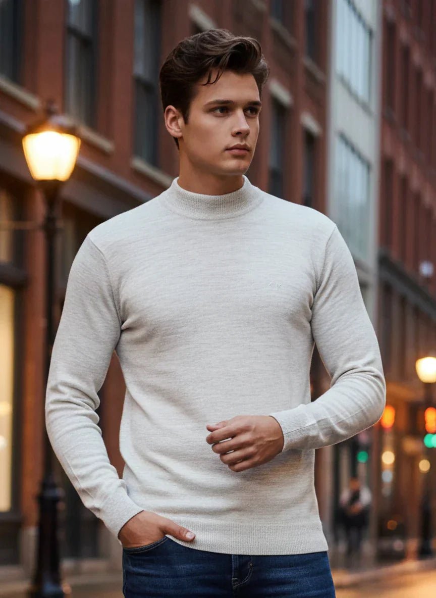 A man wears the 98degreenorth Men’s Ecru Merino Wool Turtle Neck Sweater on a city street with blurred lights behind him.