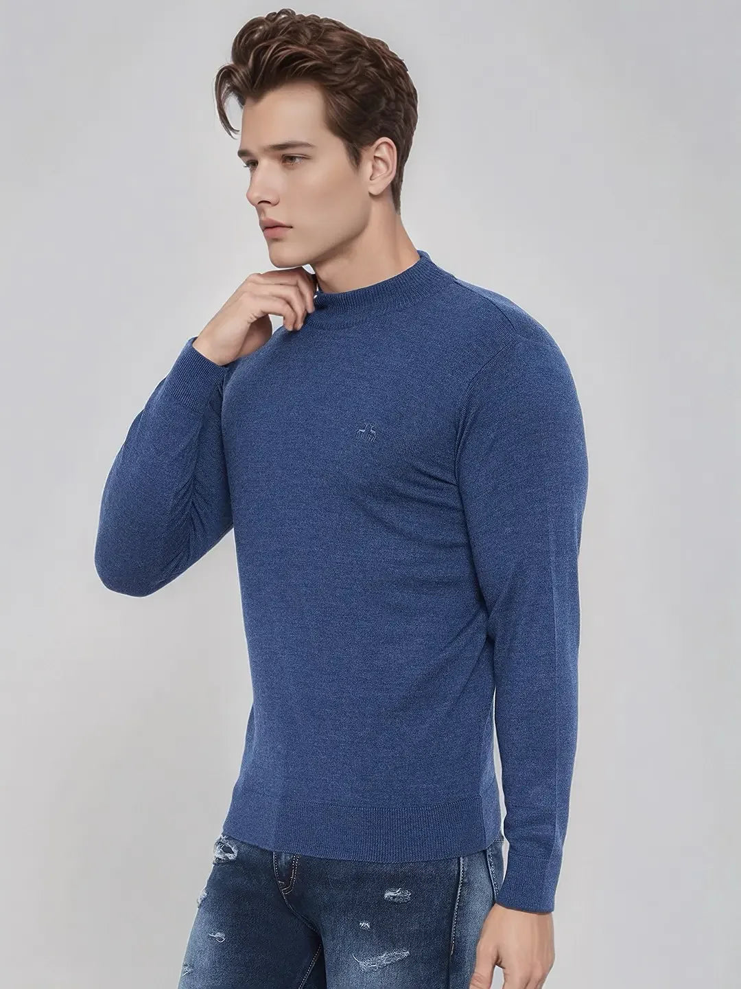A man in a 98degreenorth men’s light denim merino wool turtle neck sweater and ripped jeans stands by a plain background.