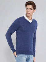 Man wears 98degreenorth Men’s Light Denim Merino Wool V-Neck Sweater over a white shirt and jeans, against plain backdrop.