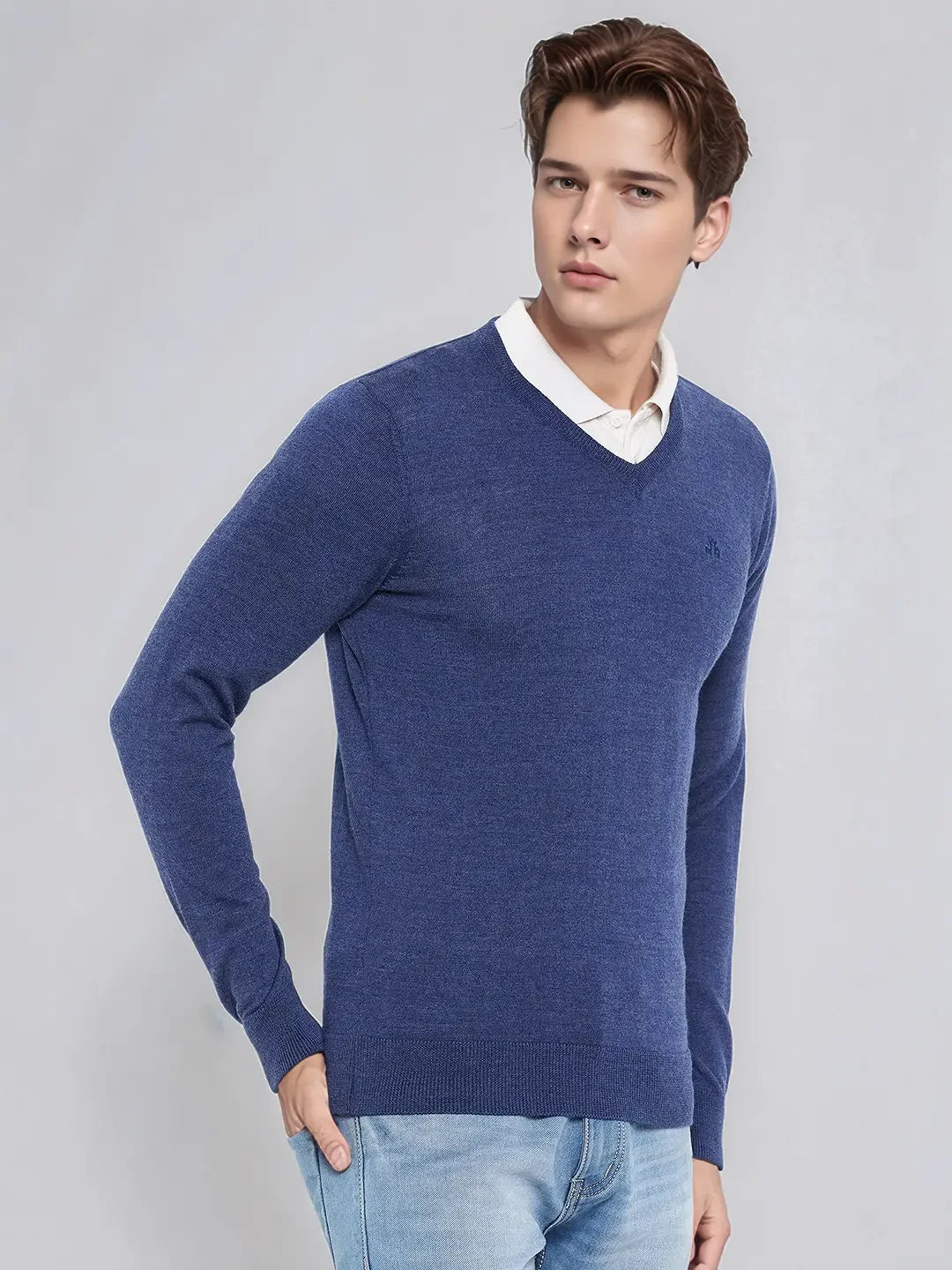 Man wears 98degreenorth Men’s Light Denim Merino Wool V-Neck Sweater over a white shirt and jeans, against plain backdrop.