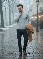 A man in a 98degreenorth Light Grey Merino Wool Turtle Neck walks a wet, foggy street with a tan coat, lights aglow behind.