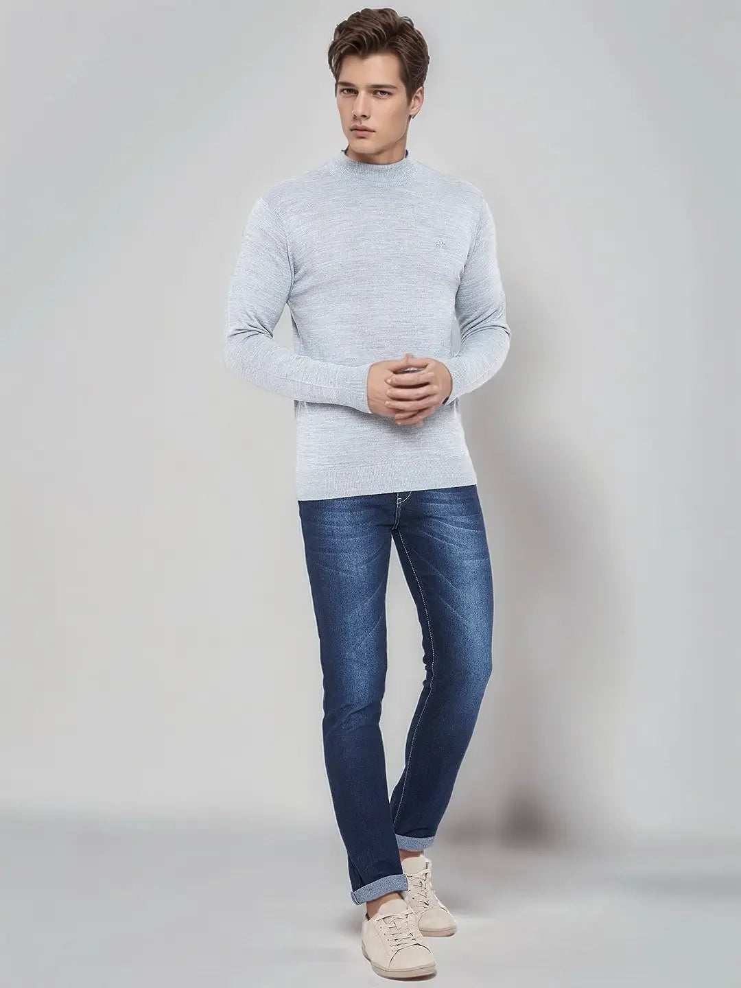 A man in a 98degreenorth light grey merino wool turtleneck sweater, blue jeans, and beige sneakers stands indoors.