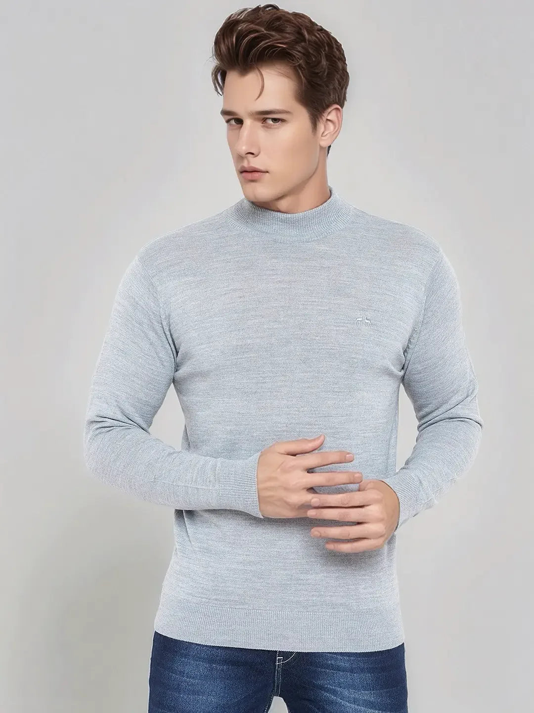 A man wears a 98degreenorth light grey merino wool turtle neck sweater and blue jeans against a plain light background.