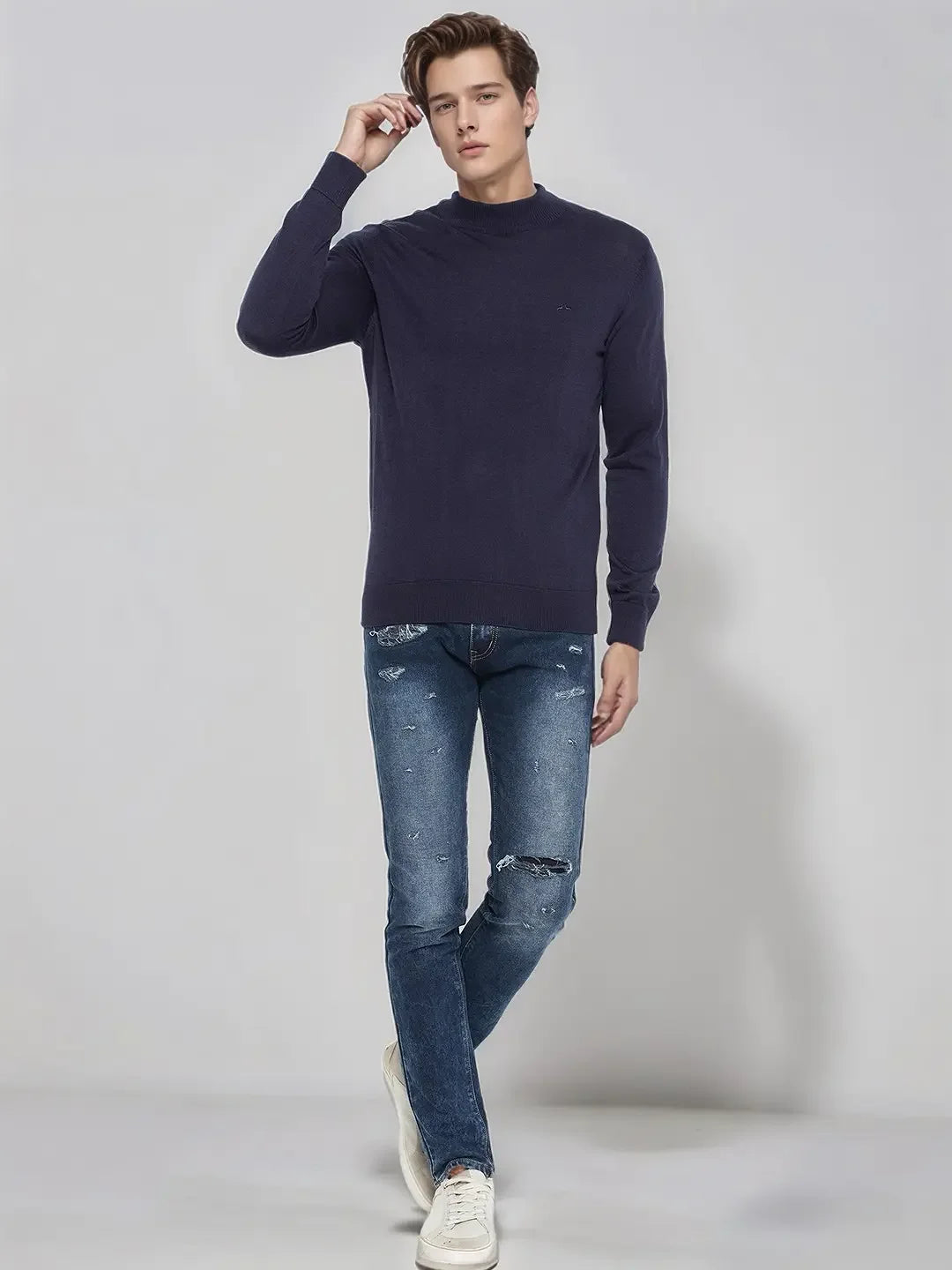 A man in a 98degreenorth light navy merino wool turtle neck sweater, ripped jeans, and white sneakers poses.