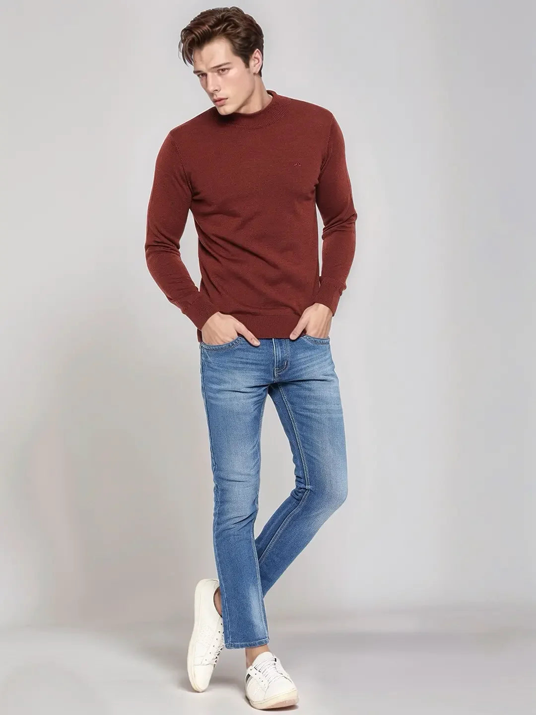 Man in a 98degreenorth Men’s Maroon Merino Wool Turtle Neck Sweater, blue jeans & white sneakers, standing against light bg.