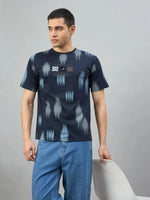 Man wears 98degreenorth Men's Navy Abstract Ikat Print T-Shirt and blue jeans, standing by a white chair against gray.