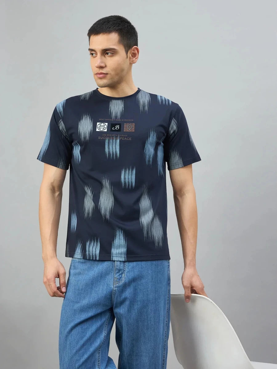 Man wears 98degreenorth Men's Navy Abstract Ikat Print T-Shirt and blue jeans, standing by a white chair against gray.