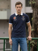 Man in a Men's Navy Aeronorth KC-767A Aviation Polo T-Shirt by 98degreenorth stands outdoors by a green railing.