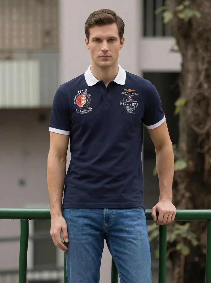 Man in a Men's Navy Aeronorth KC-767A Aviation Polo T-Shirt by 98degreenorth stands outdoors by a green railing.
