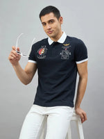 Man wears 98degreenorth Men's Navy Aeronorth KC-767A Aviation Polo T-Shirt, holding glasses on a stool against gray.