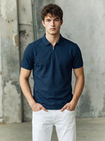 A man in a 98degreenorth navy blue popcorn knit quarter zip polo and white pants stands against a gray industrial wall.