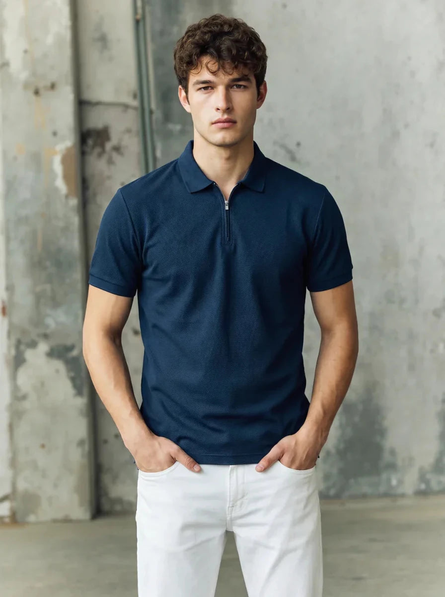 A man in a 98degreenorth navy blue popcorn knit quarter zip polo and white pants stands against a gray industrial wall.