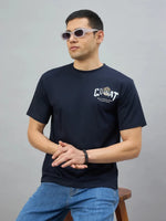 Man in sunglasses wears a navy oversized 98degreenorth GOAT Back Print T-shirt and blue jeans, sitting on a stool.