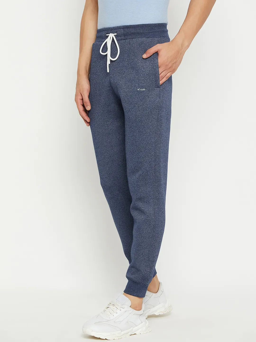 A person in 98degreenorth Men’s Navy Jaspe Fleece Trackpants, a light blue shirt, and white sneakers stands against a plain background.