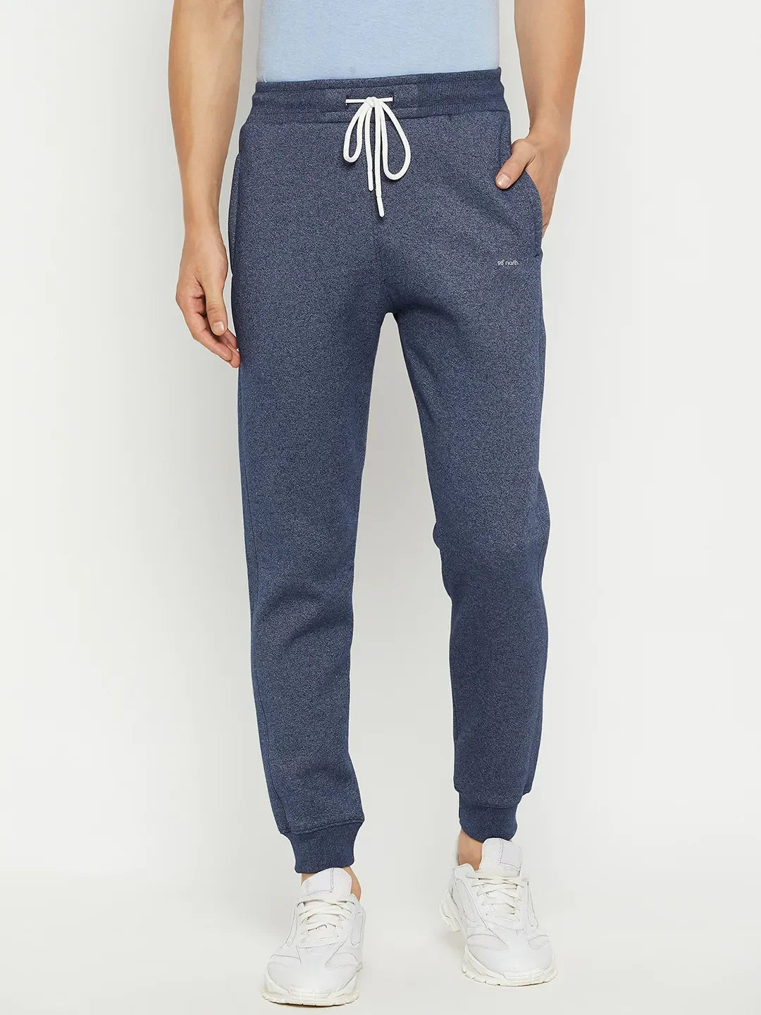 Man wears 98degreenorth Men's Navy Jaspe Fleece Trackpants, a light blue shirt, and white sneakers against a white background.