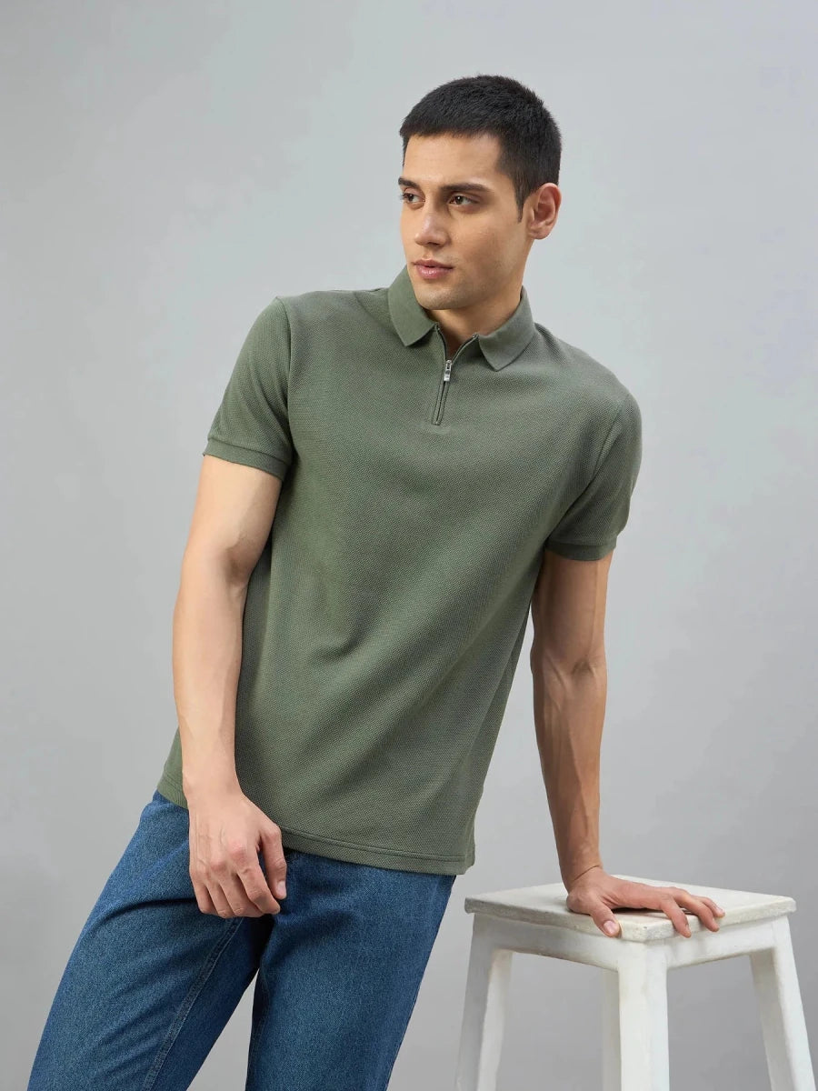 Man wears 98degreenorth Olive Green Popcorn Knit Quarter Zip Polo T-Shirt with blue jeans, by a white stool on gray.