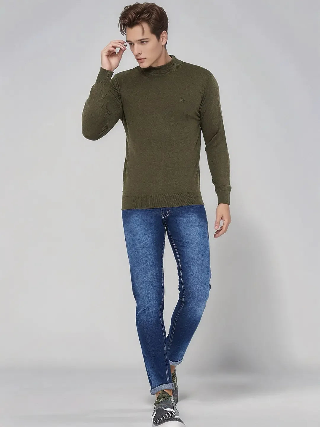 Man in 98degreenorth Men’s Olive Merino Wool Turtle Neck Sweater, blue jeans & sneakers, walks against light gray background.