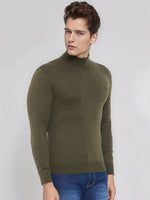 A man wears the 98degreenorth Men’s Olive Merino Wool Turtle Neck Sweater with blue jeans against a light gray background.