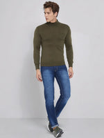 A man wears the 98degreenorth Olive Merino Wool Turtle Neck Sweater with blue jeans and sneakers against a light gray backdrop.
