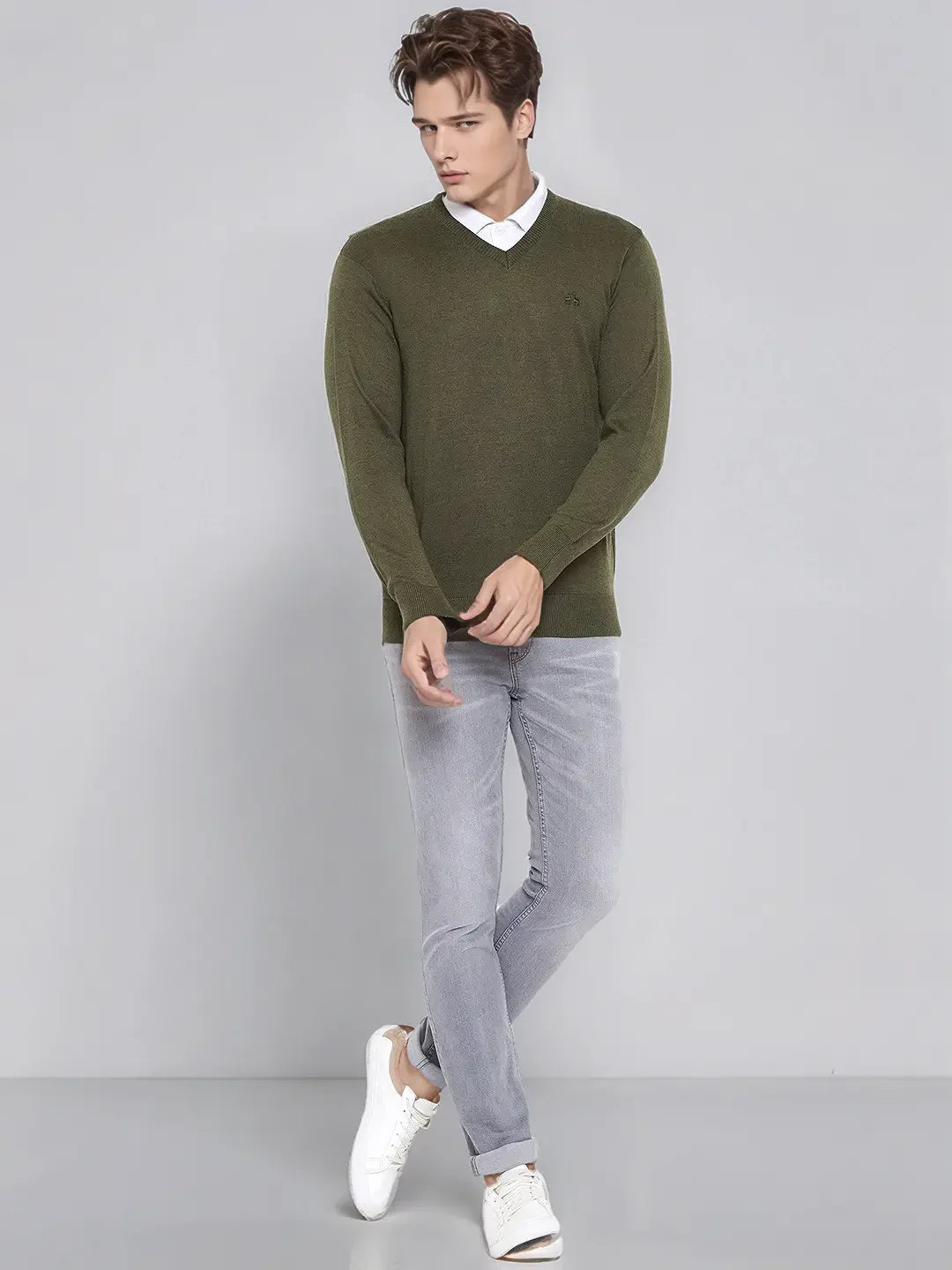Man wears 98degreenorth Men’s Olive Merino Wool V-Neck Sweater, gray jeans, and white sneakers against a gray background.