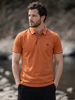Man in a 98degreenorth Men's Orange Polo T-Shirt, Handmade Series with contrast tipping, stands by rocks and water.