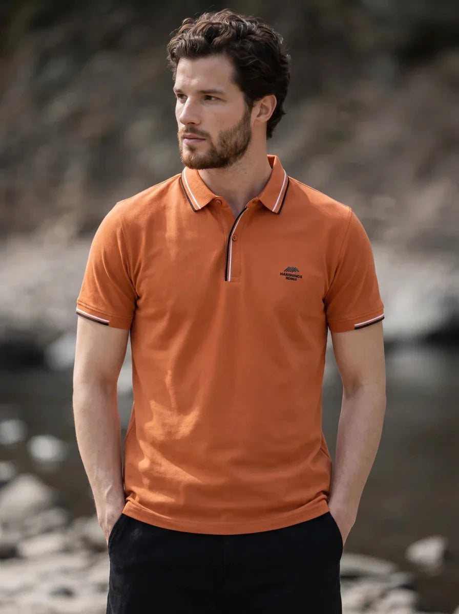Man in a 98degreenorth Men's Orange Polo T-Shirt, Handmade Series with contrast tipping, stands by rocks and water.