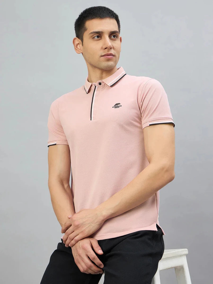 A man in a 98degreenorth Men's Pink Polo T-Shirt sits on a white stool against a plain grey background.