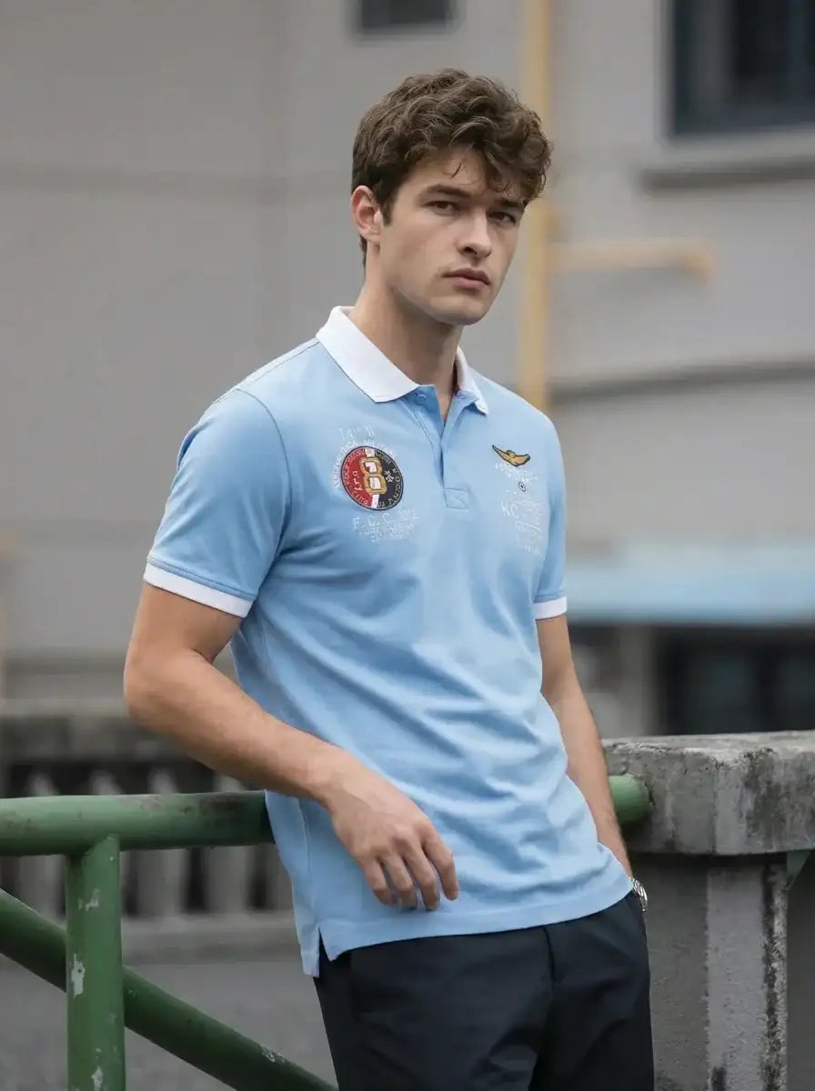 Young man in a 98degreenorth Sky Blue KC-767A Aviation Polo T-Shirt leans on a railing, looking at the camera.