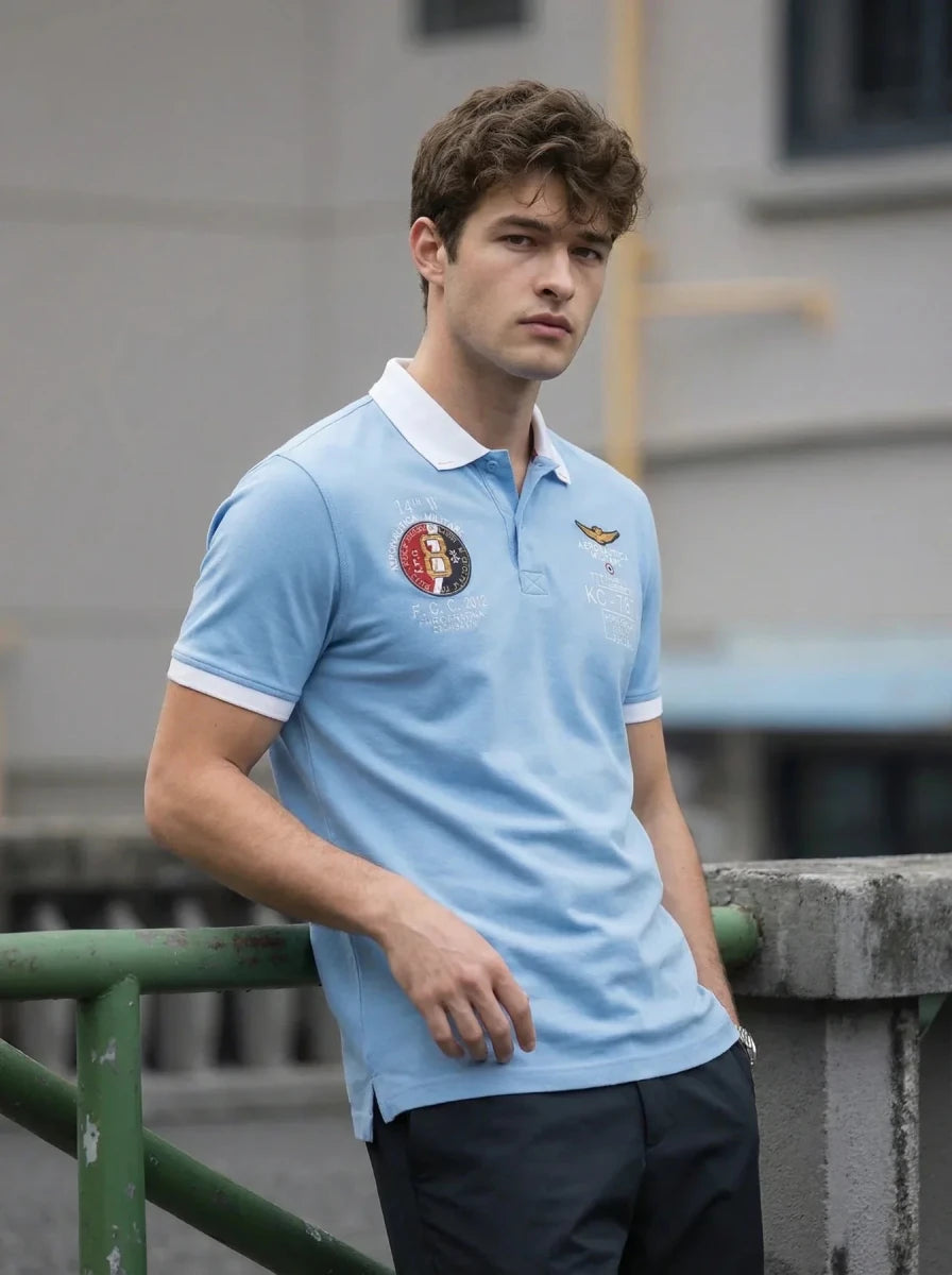 Young man in a 98degreenorth Sky Blue KC-767A Aviation Polo T-Shirt leans on a railing, looking at the camera.