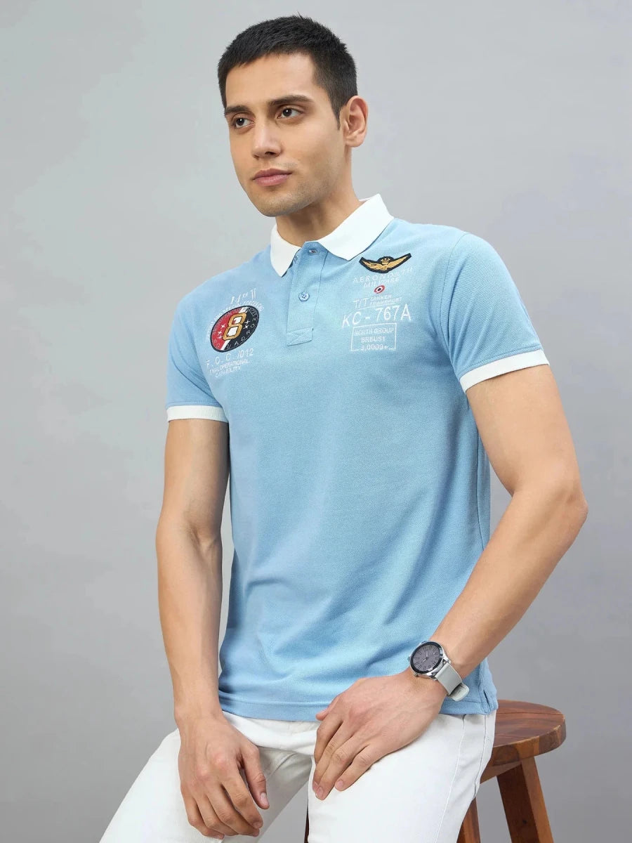 Man in a Men's Sky Blue Aeronorth KC-767A Aviation Polo T-Shirt by 98degreenorth sits on a stool against a gray background.
