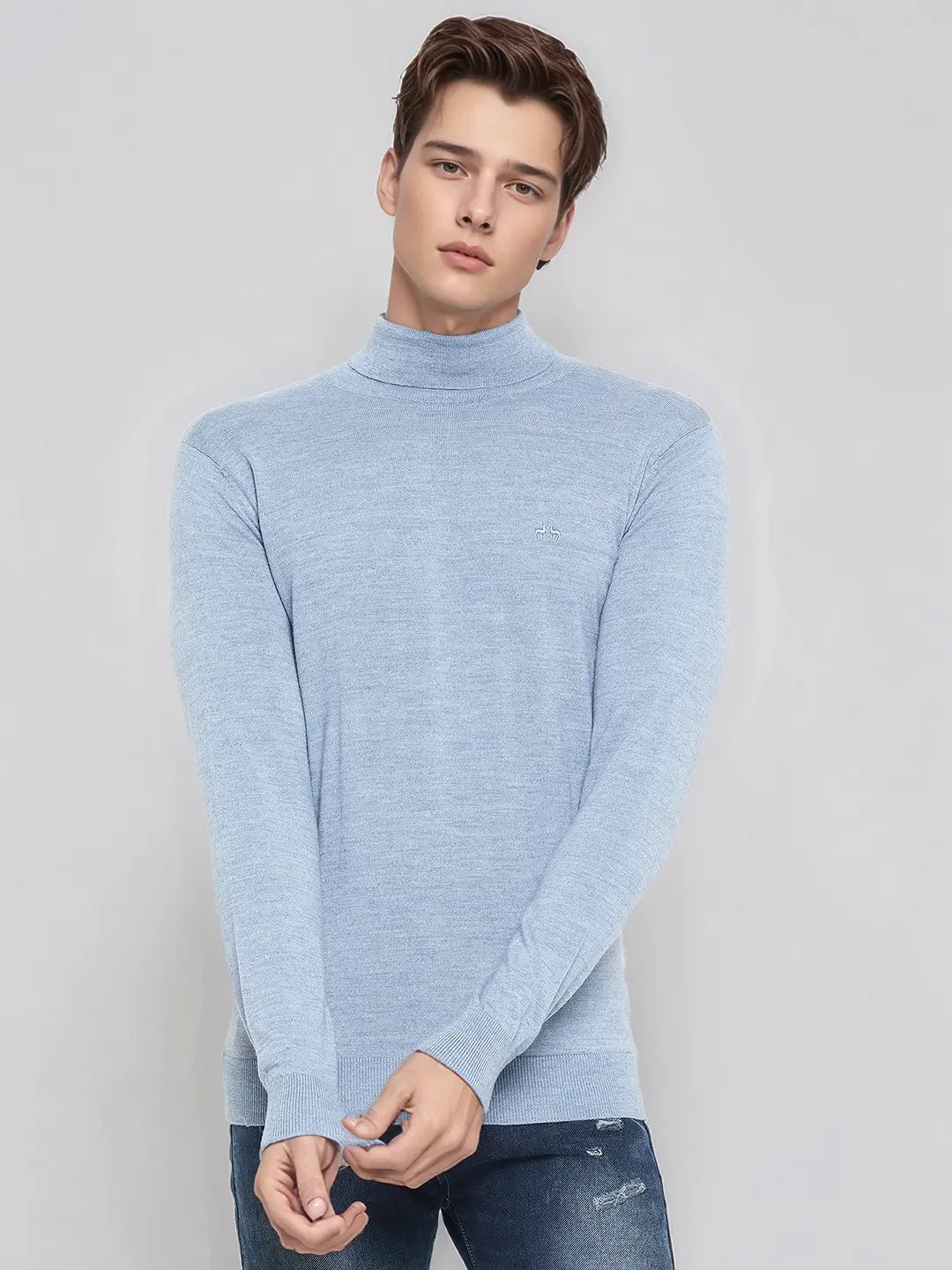 A young man in a 98degreenorth sky blue merino wool turtleneck sweater and jeans stands against a light background.