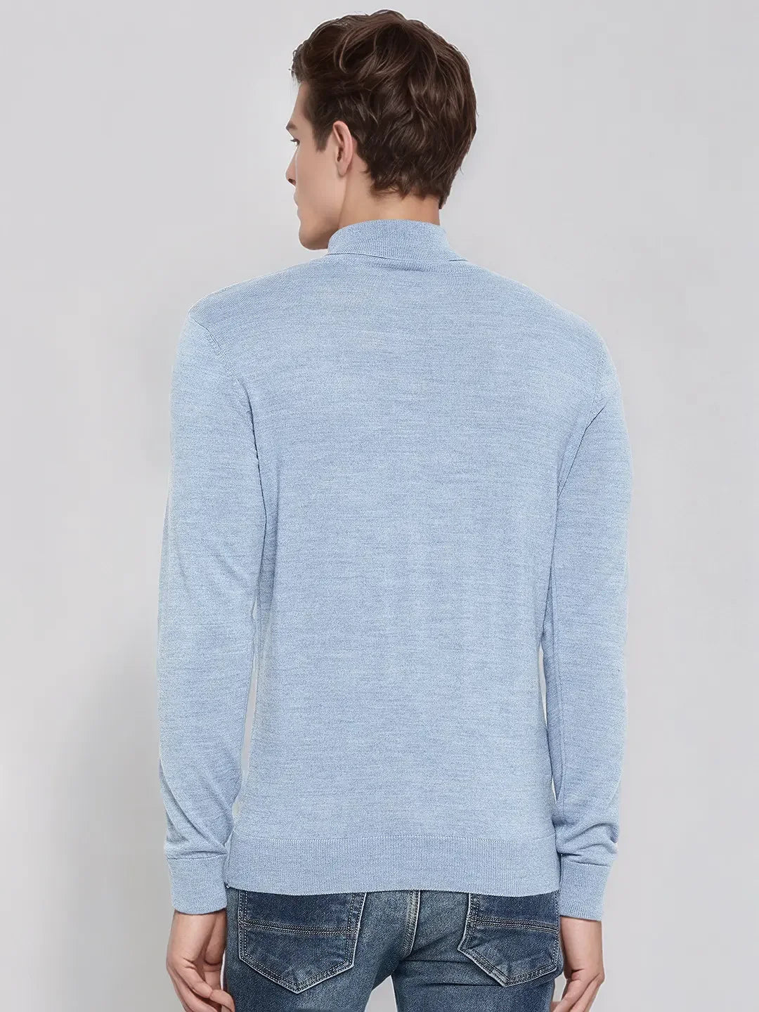 A man in a 98degreenorth sky blue merino wool turtleneck sweater and jeans stands facing away against a plain background.