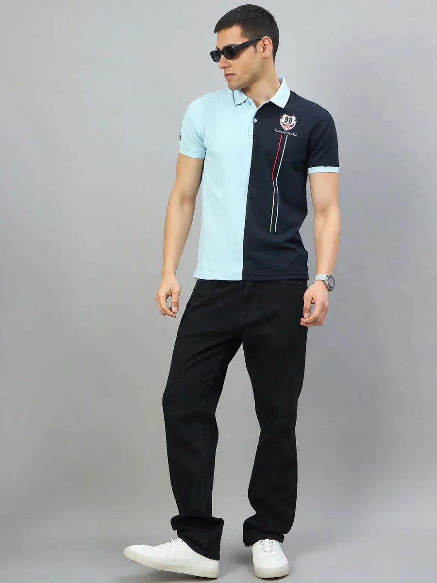 Man in black jeans and a sky blue & navy colour block polo by 98degreenorth stands against a gray background.