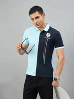 A man in a 98degreenorth sky blue & navy colour block polo stands by a white chair against a gray background.
