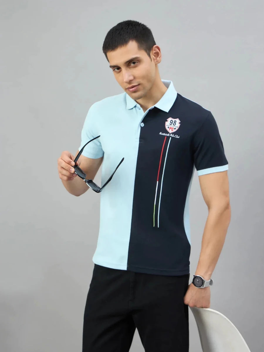 A man in a 98degreenorth sky blue & navy colour block polo stands by a white chair against a gray background.