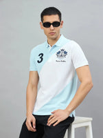 Man in 98degreenorth Men's Sky Blue & White Colour Block Polo T-Shirt with crest, sitting on a stool.