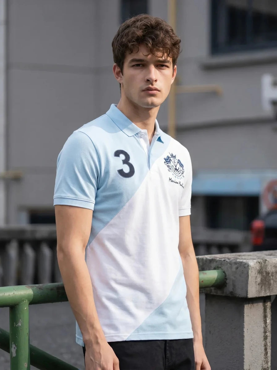 A man wears a 98degreenorth Sky Blue & White Colour Block Polo with Marine Harbns crest, standing outdoors by a railing.