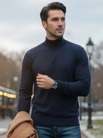 A man in a 98degreenorth teal blue Merino wool turtle neck stands outdoors holding a brown coat under streetlights.
