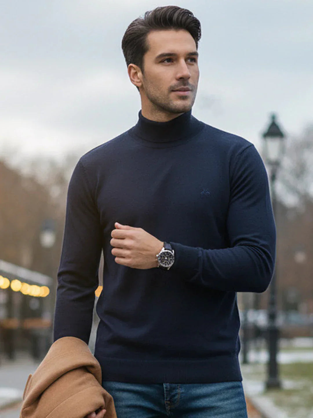 A man in a 98degreenorth teal blue Merino wool turtle neck stands outdoors holding a brown coat under streetlights.